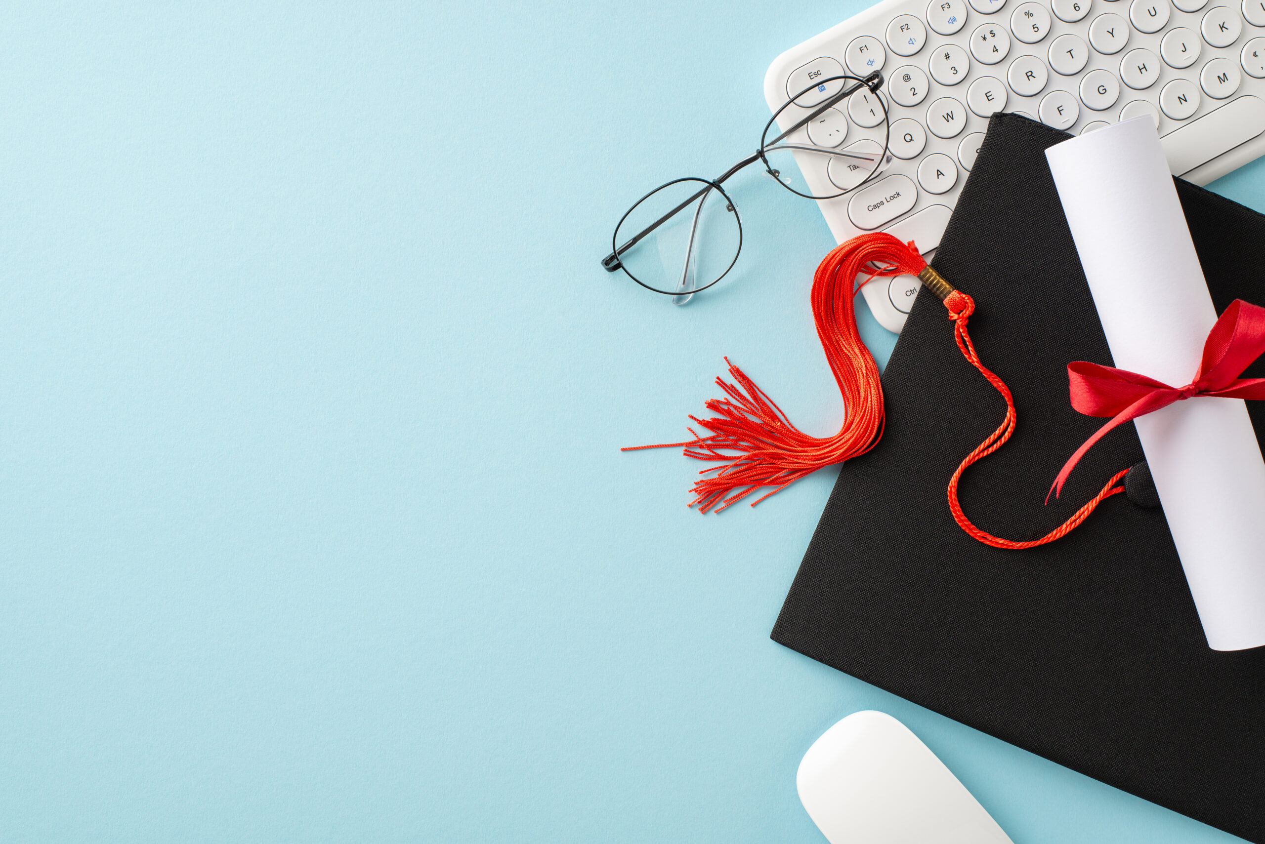 top view of graduation cap alongside a diploma, tied with a red ribbon, white keyboard